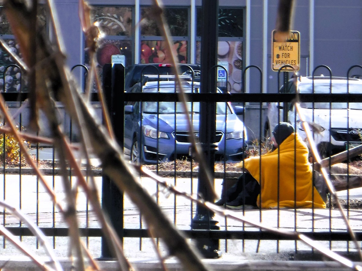 A person sitting wrapped in a yellow blanket in front of a parked car.