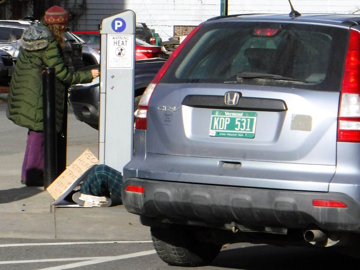A person paying at a parking meter next to a silver car. There are knees and shoes visible with a sign leaning on them asking for money.