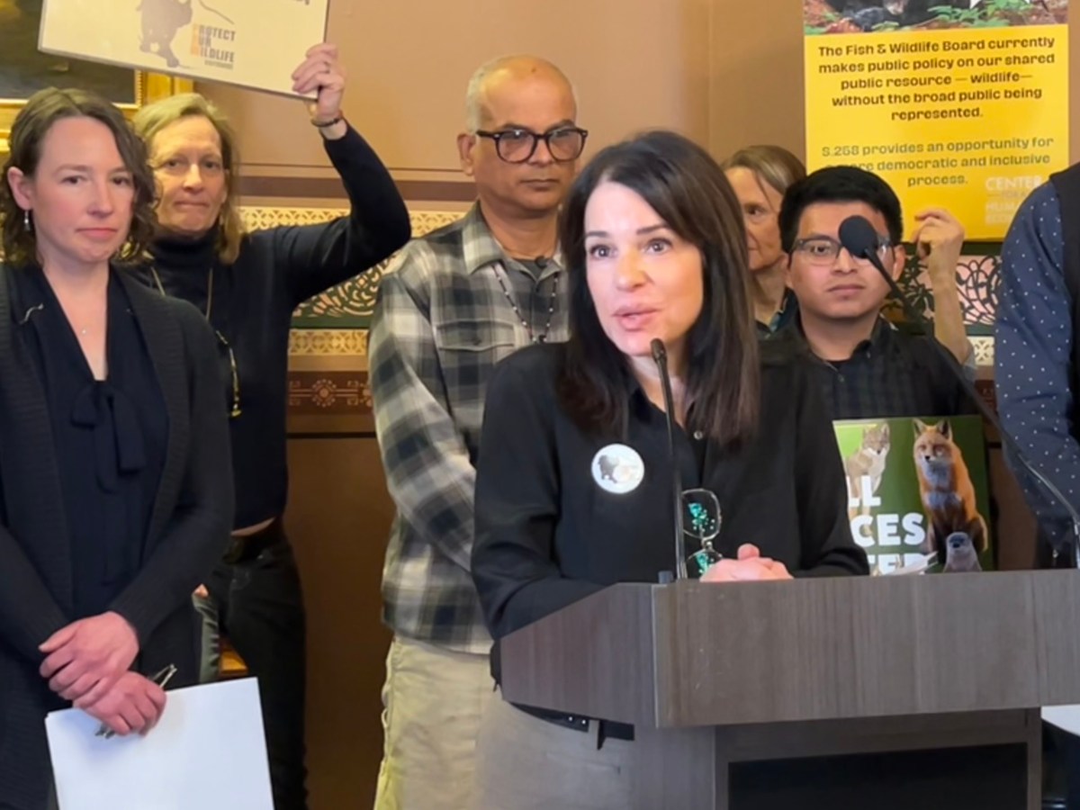 Woman speaking at a podium during a public event, with several attentive people standing behind her in a room with posters.