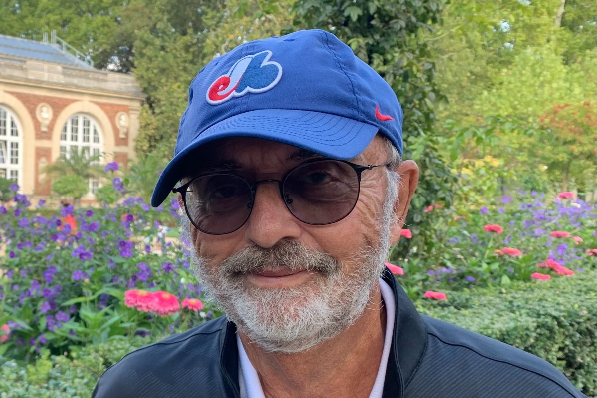 A man with a gray beard and glasses, wearing a blue cap with a red and white logo, stands in a garden with colorful flowers and a building in the background.