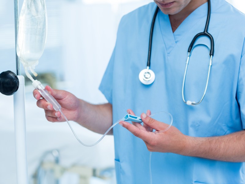 A doctor in light blue scrubs and a stethoscope around his neck examines an intravenous drip.