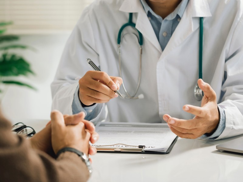 A doctor in a white coat and stethoscope talks to a patient.