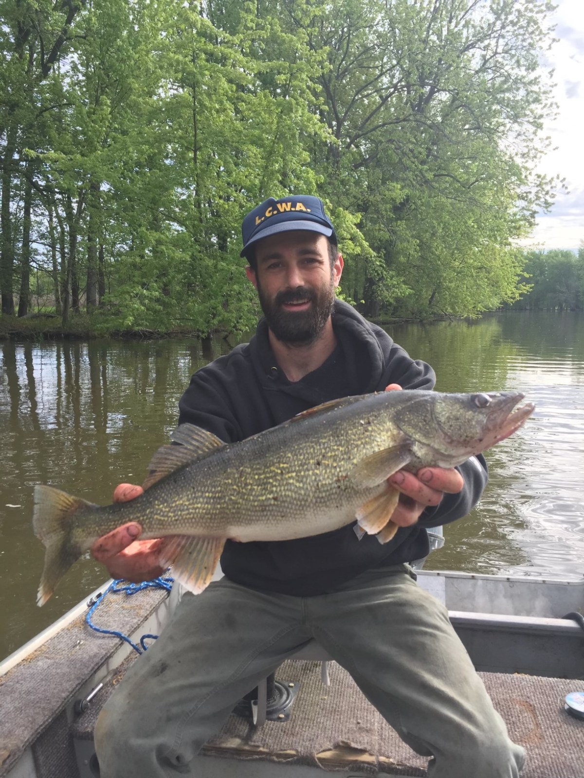 A man in a boat holding a large fish with both hands, surrounded by trees and water.