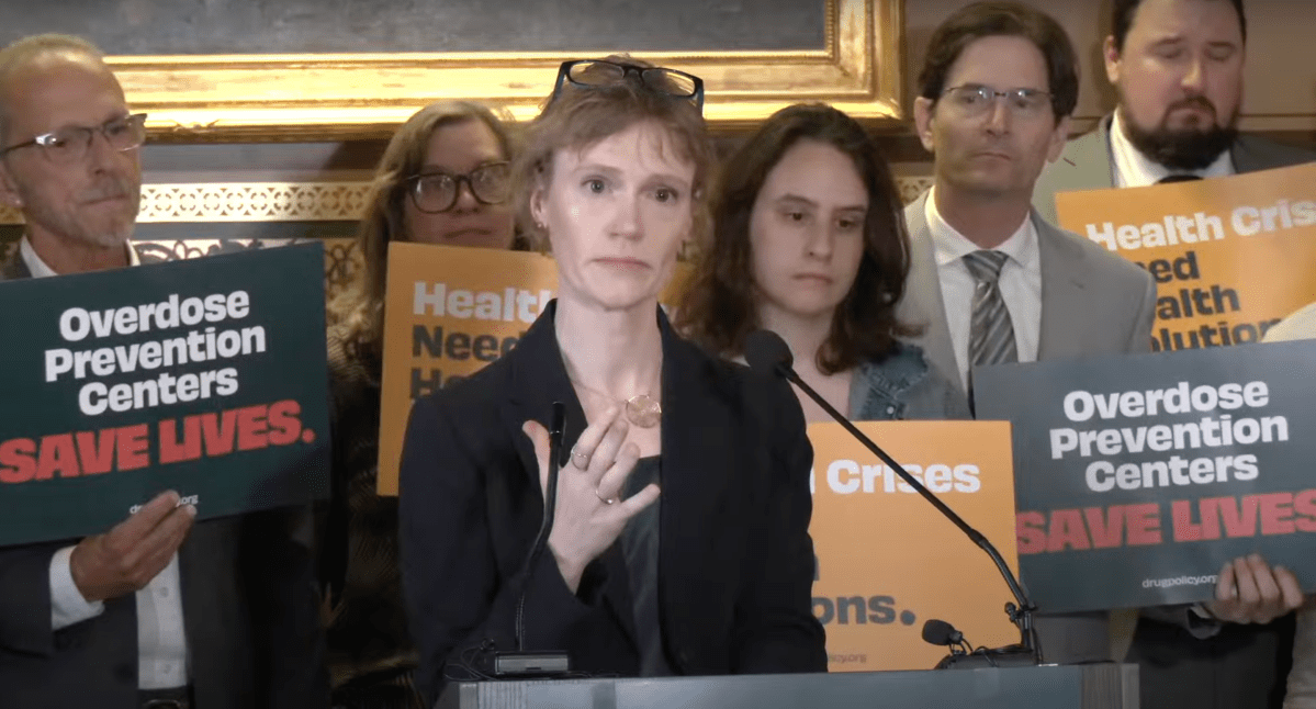 Woman speaking at a podium with "overdose prevention centers save lives" signs held by attendees in the background.