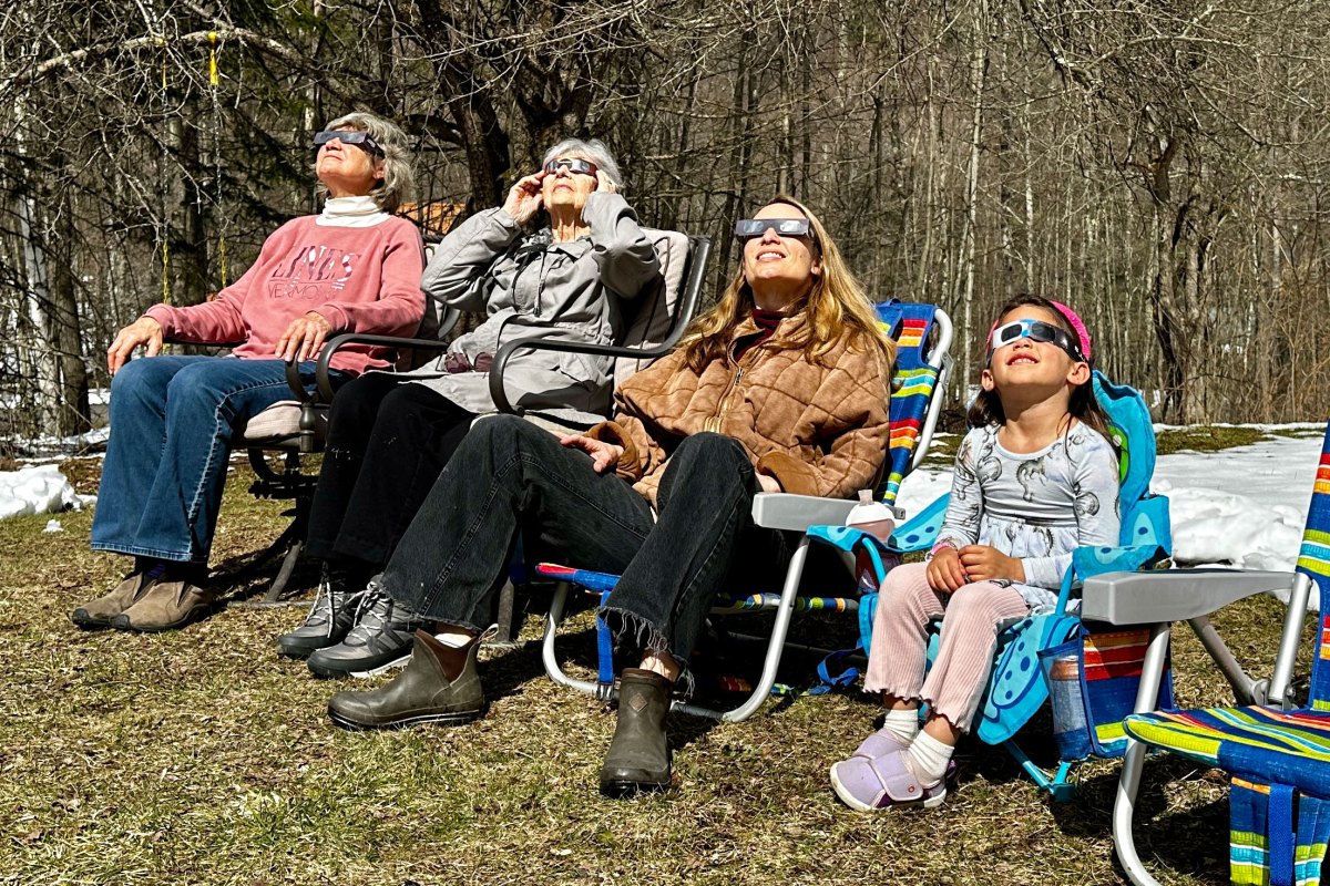 Three women and one girl sit in lawn chairs and look up at the sky while wearing eclipse glasses.