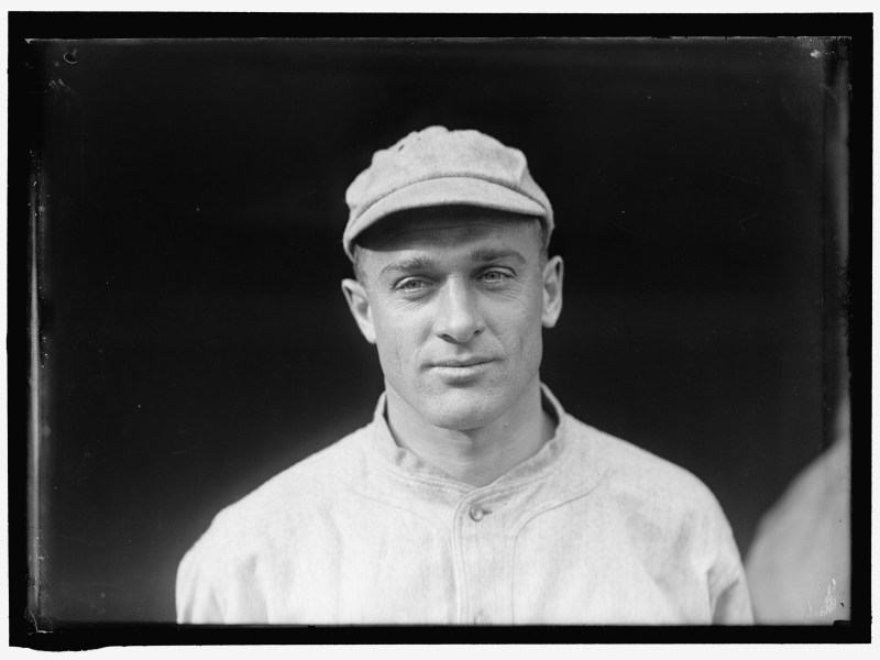 A black and white photograph of a man wearing a cap and a buttoned shirt, looking at the camera with a neutral expression.