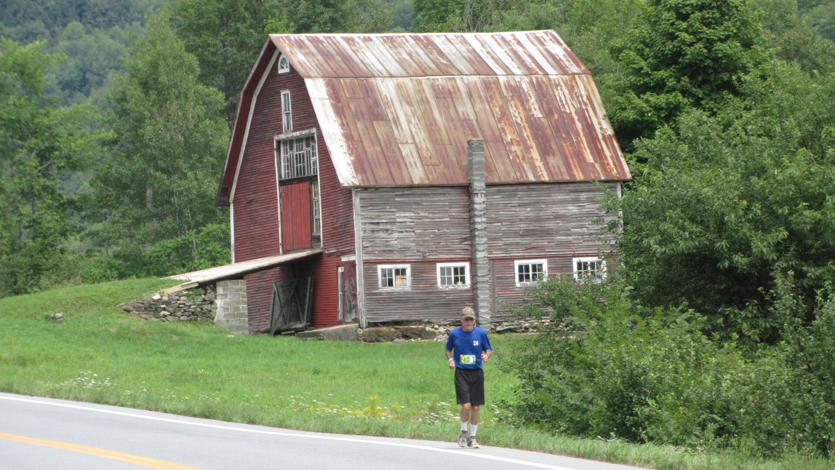 Man running down a road by an old red barn
