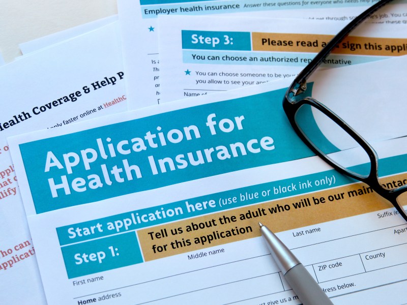 A small pile of papers for an application for health insurance with a pair of glasses and a pen on top.