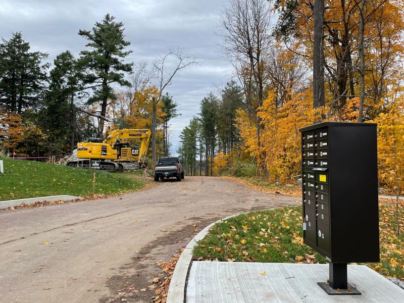 A construction site with a yellow excavator and a black truck on a dirt road amidst autumn trees. A black mailbox unit is visible in the foreground.