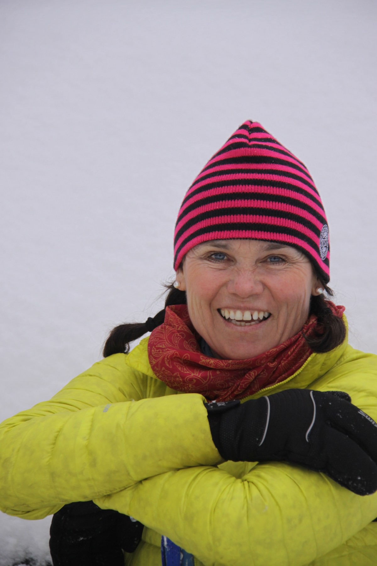 A smiling woman in a striped hat and yellow jacket standing against a snowy backdrop.