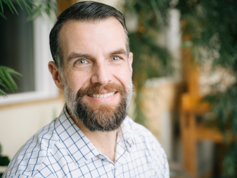 A man with a beard smiling in front of a plant.