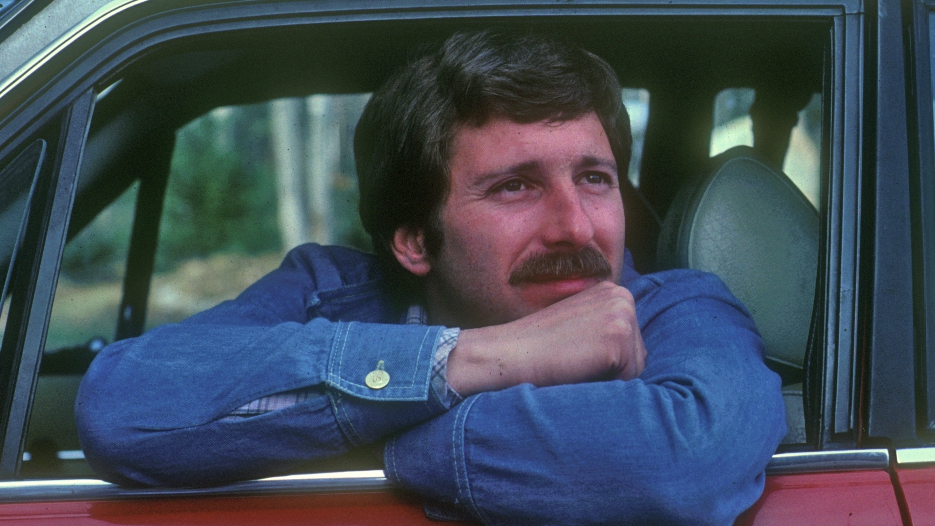 A man with a mustache leaning out of a red car window, resting his chin on his arm, wearing a denim jacket.