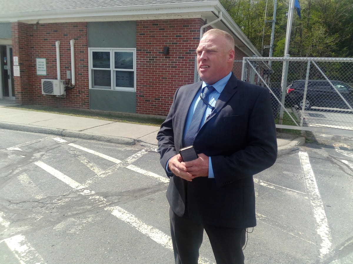 A man in a suit stands outside a brick building with a smartphone in his hand, near a chain-link fence and parked cars.