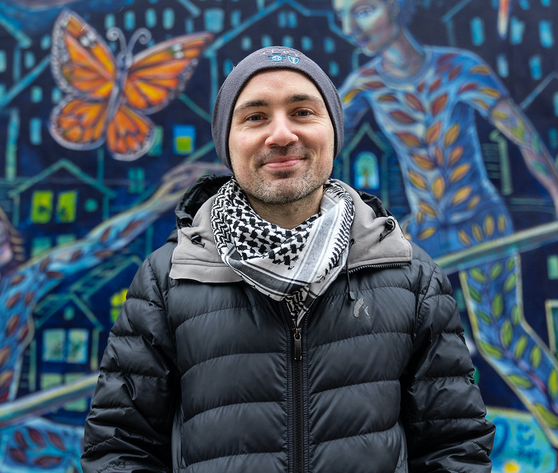 A man in a black coat and beanie standing in front of a mural.