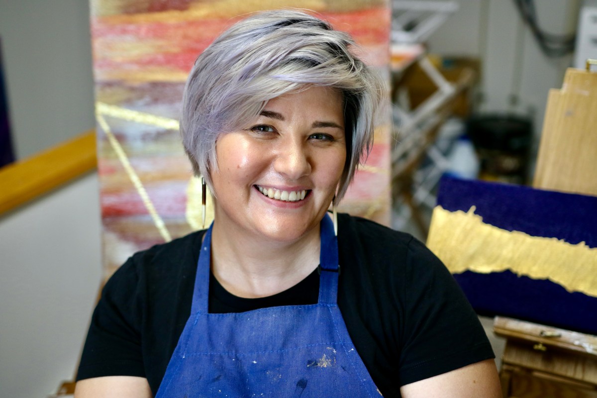 A woman in an apron smiling in front of a painting.
