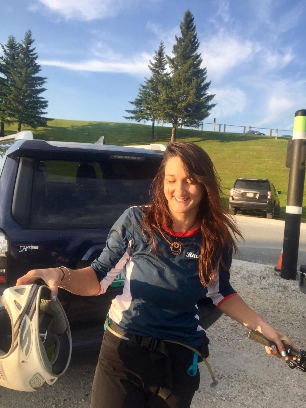 A woman smiles while holding a bicycle helmet next to a parked suv in a sunny parking area with trees in the background.