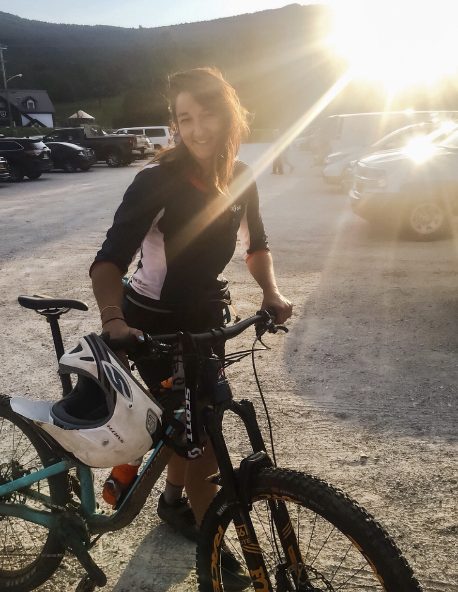 A smiling woman stands next to a mountain bike in a parking lot, with the sun setting in the background.