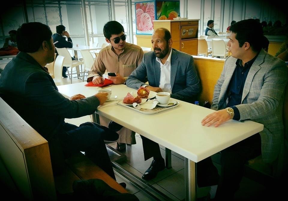 Four men sit around a white table in a cafeteria, engaging in conversation. One tray with food and drinks is on the table. They are dressed in semi-formal attire.