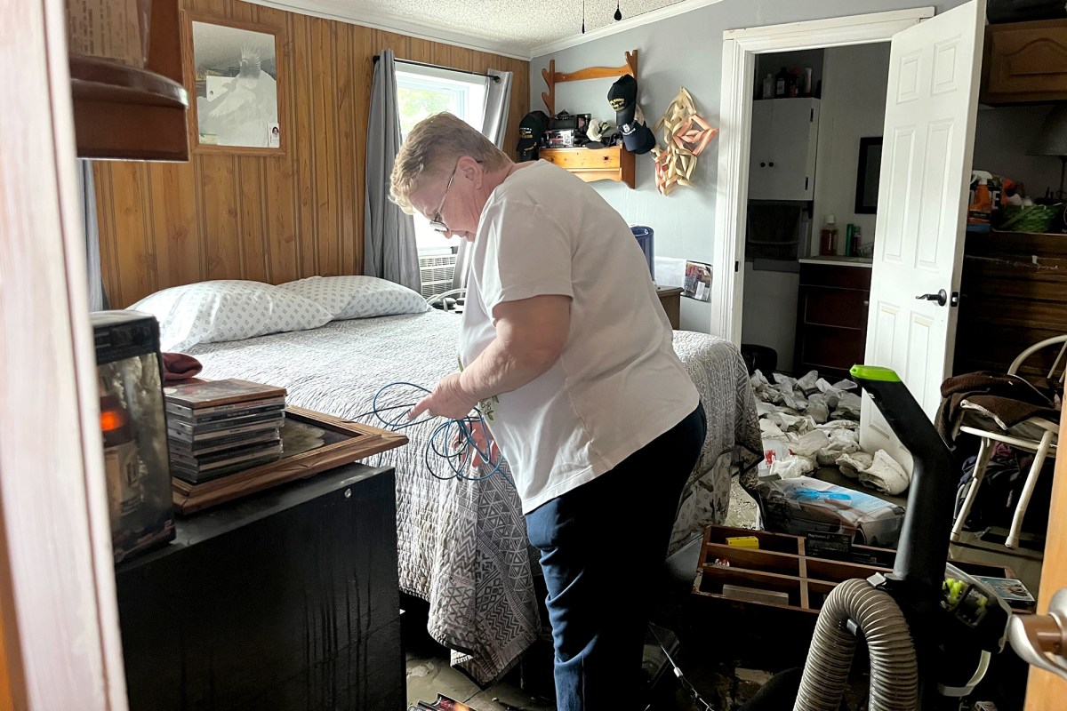 A woman in a white shirt stands in front of a bed.