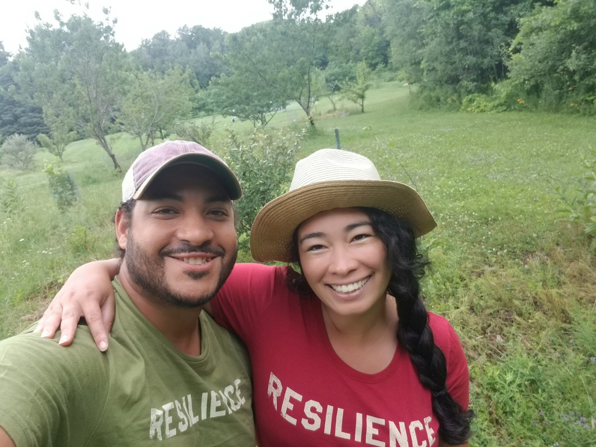 A smiling man and woman wearing "RESILIENCE" shirts stand together outdoors with greenery in the background.