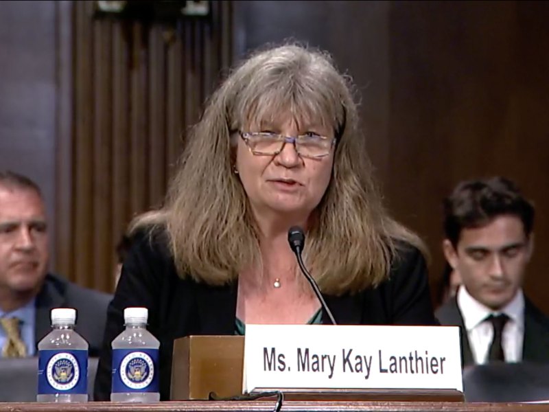 A woman wearing glasses speaks into a small microphone at a table.