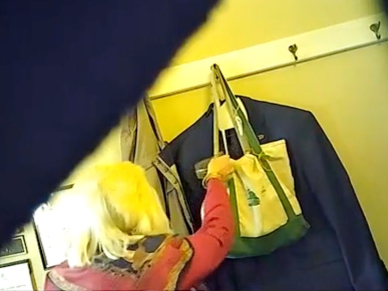 A person with light-colored hair reaches into a tote bag hanging on a wall rack with clothes, viewed through a partially obstructed angle.