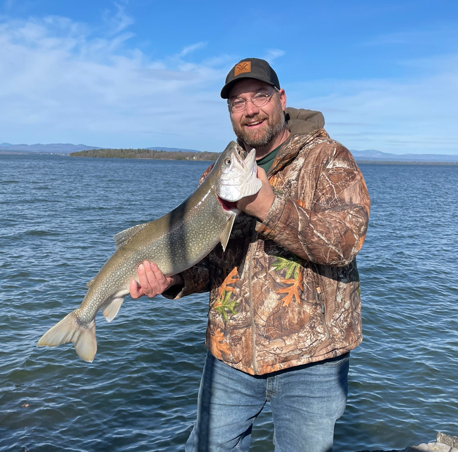 Man in camouflage jacket holding a large fish at a lakeside, smiling, with clear blue sky and distant hills in the background.