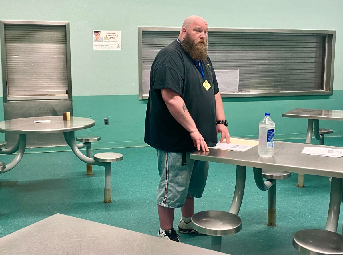 A bearded man stands by a table with papers and a water bottle in a room with metal tables and teal walls.