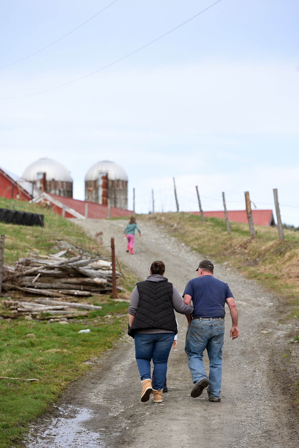 A couple walks hand in hand up a dirt road on a farm, with a child ahead and two silos in the background.