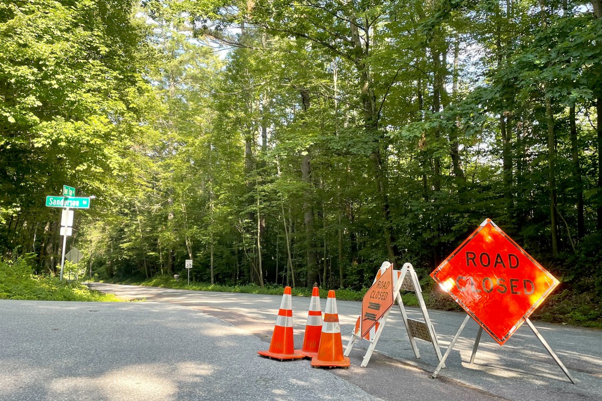 An orange "road closed" sign next to traffic cones on a street surrounded by trees.