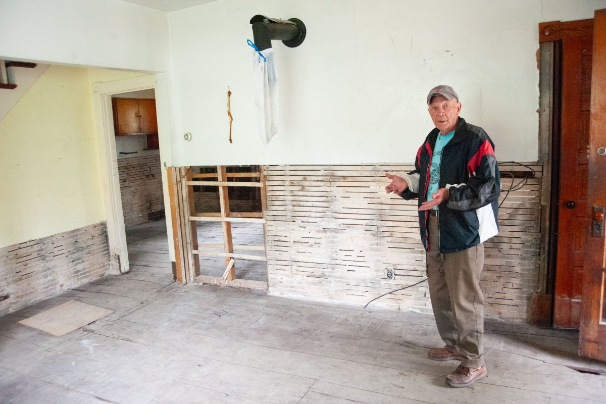 A man in a baseball camp stands in a pale yellow room that has had the bottom half of its drywall removed.