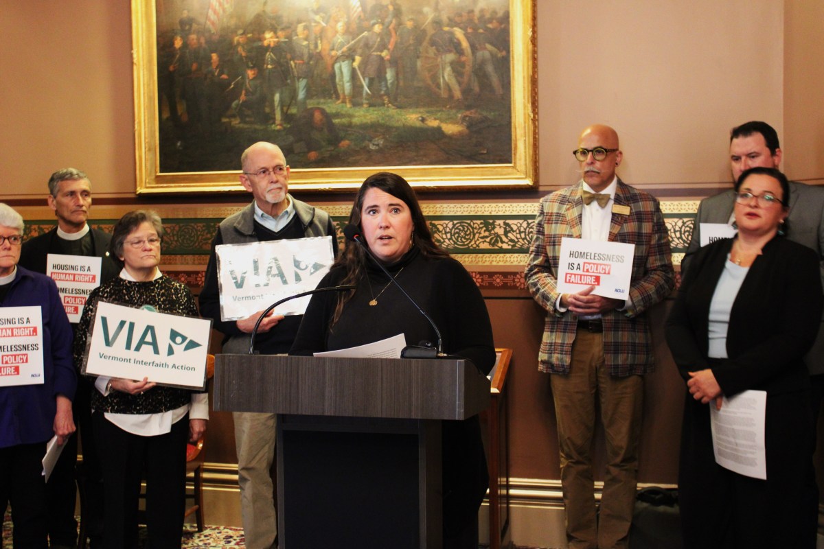 A woman speaks at a podium during a legislative event, flanked by a group of people holding signs about homelessness and healthcare.
