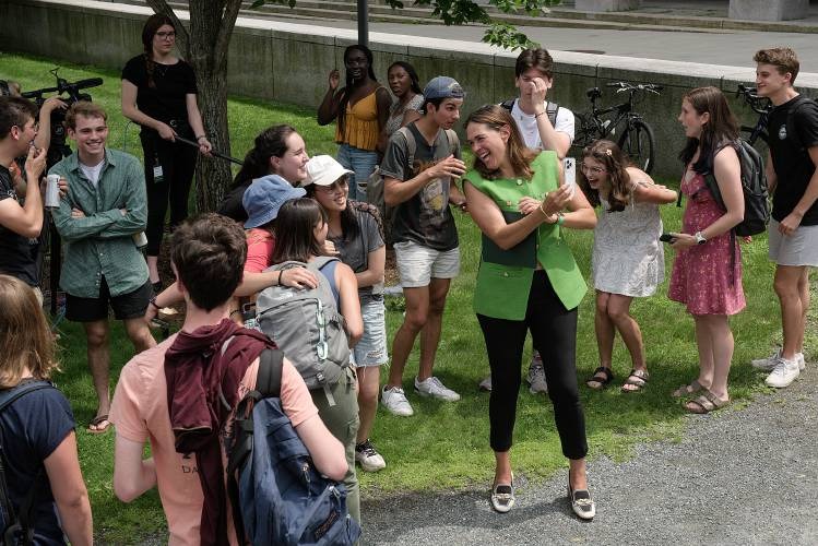 A group of young adults gather outdoors in a park, talking and laughing. A woman in a green vest is leading the group, engaging with others cheerfully. Bicycles are seen in the background.