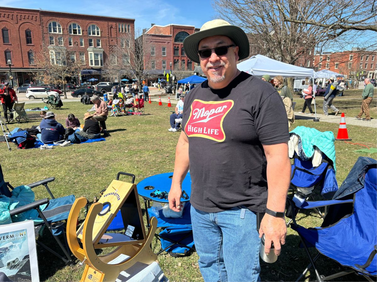 Man in a fedora and sunglasses standing at an outdoor event with booths and people on the grass in the background.