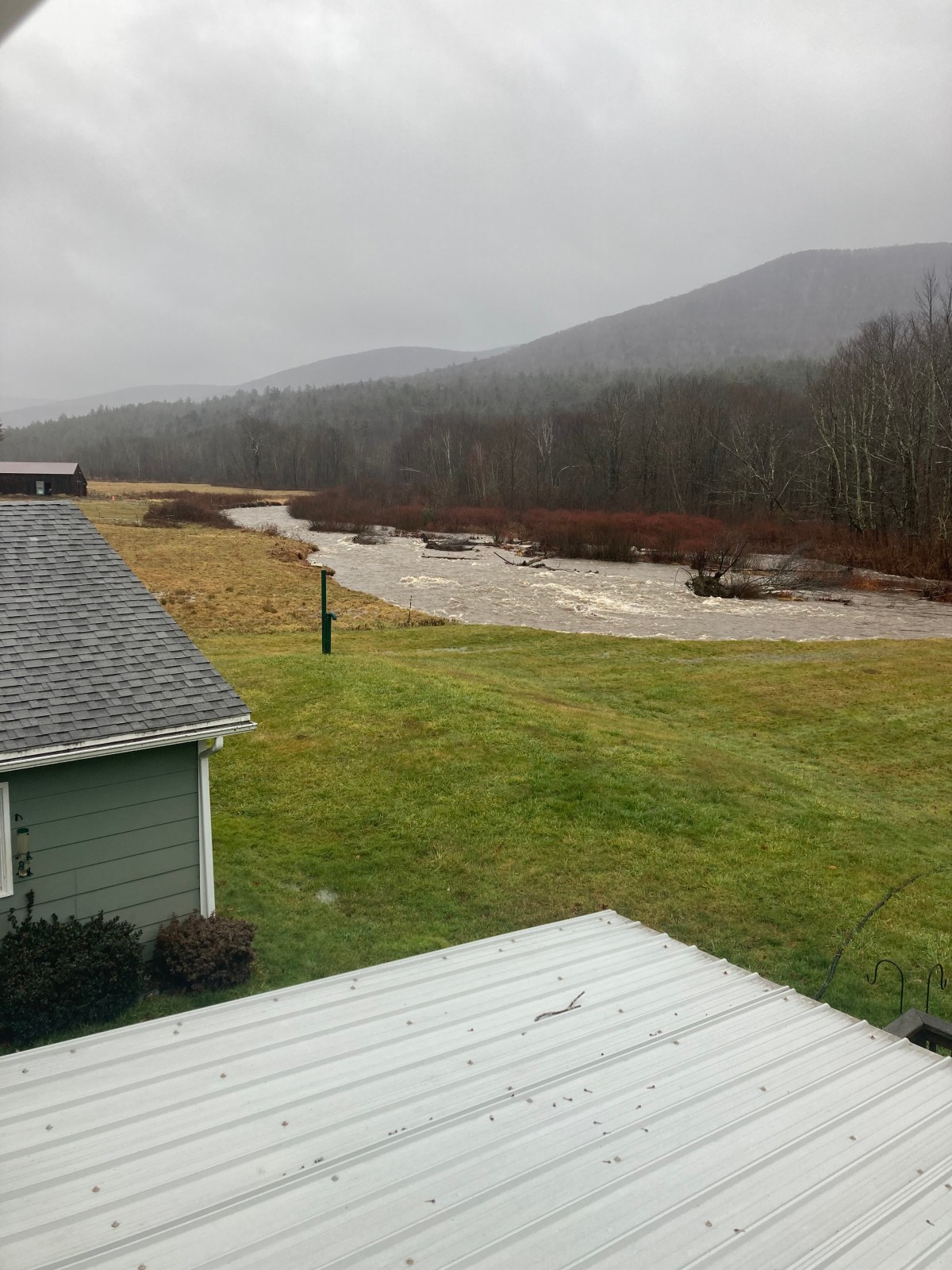 A view from above showing a swollen river beside lush green grass, with foggy mountains in the background and the corner of a building on the left.