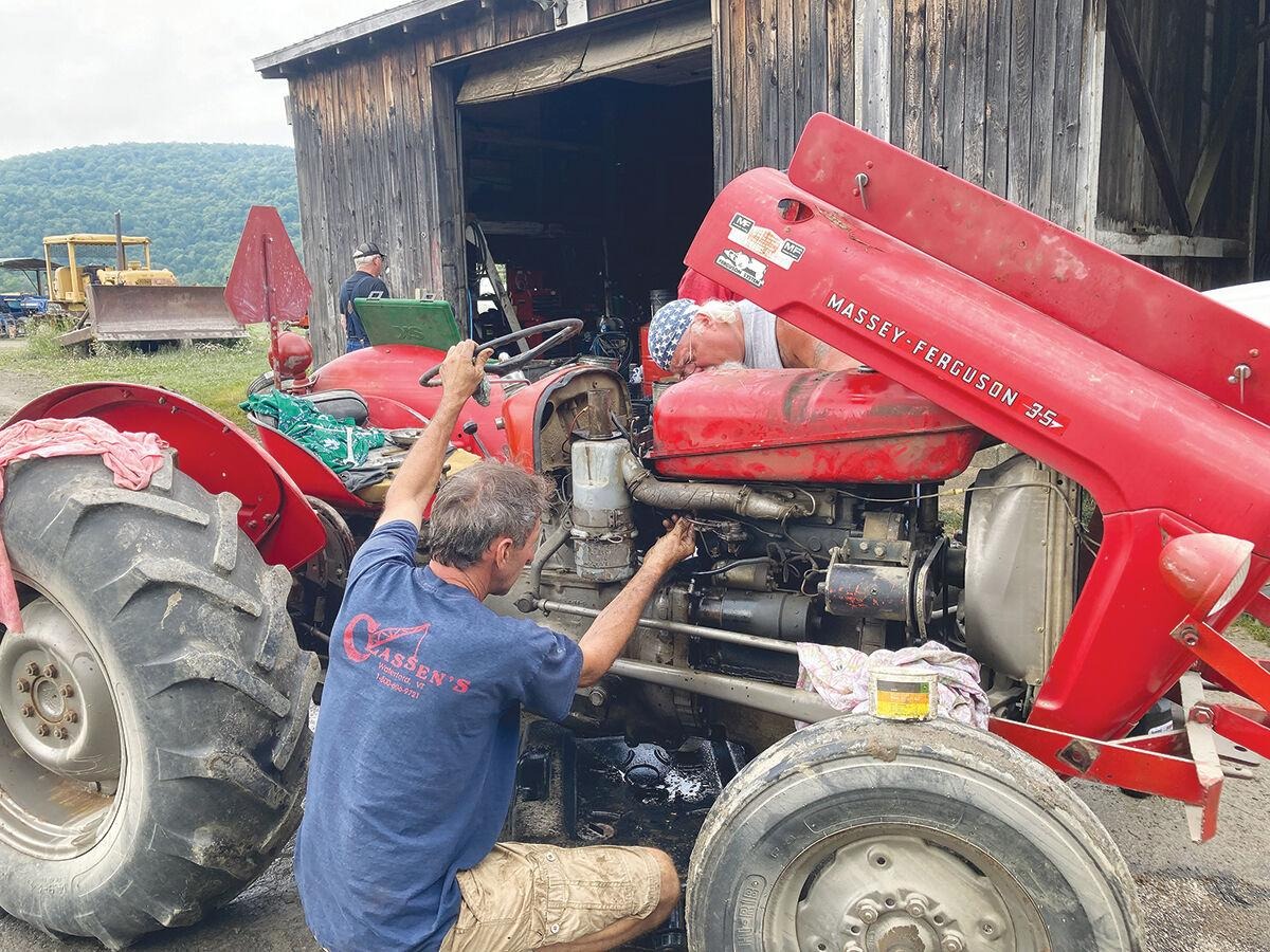 A man repairs a red Massey Ferguson 35 tractor in front of an open barn with other machinery visible inside.