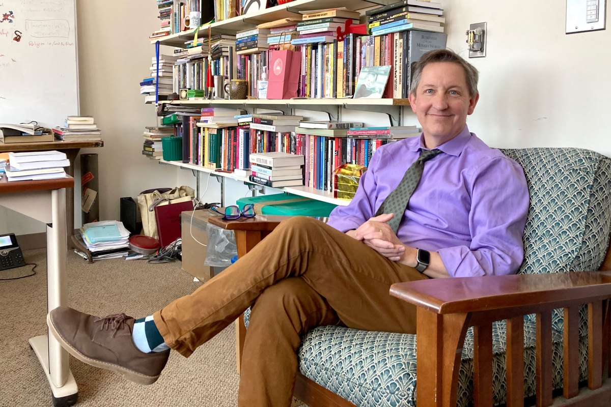 A man in a purple shirt and brown pants sits in a chair in front of bookshelves.
