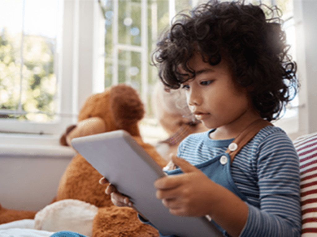 A young child with curly hair, dressed in a striped overall, intently uses a tablet while sitting near a window with a teddy bear in the background.