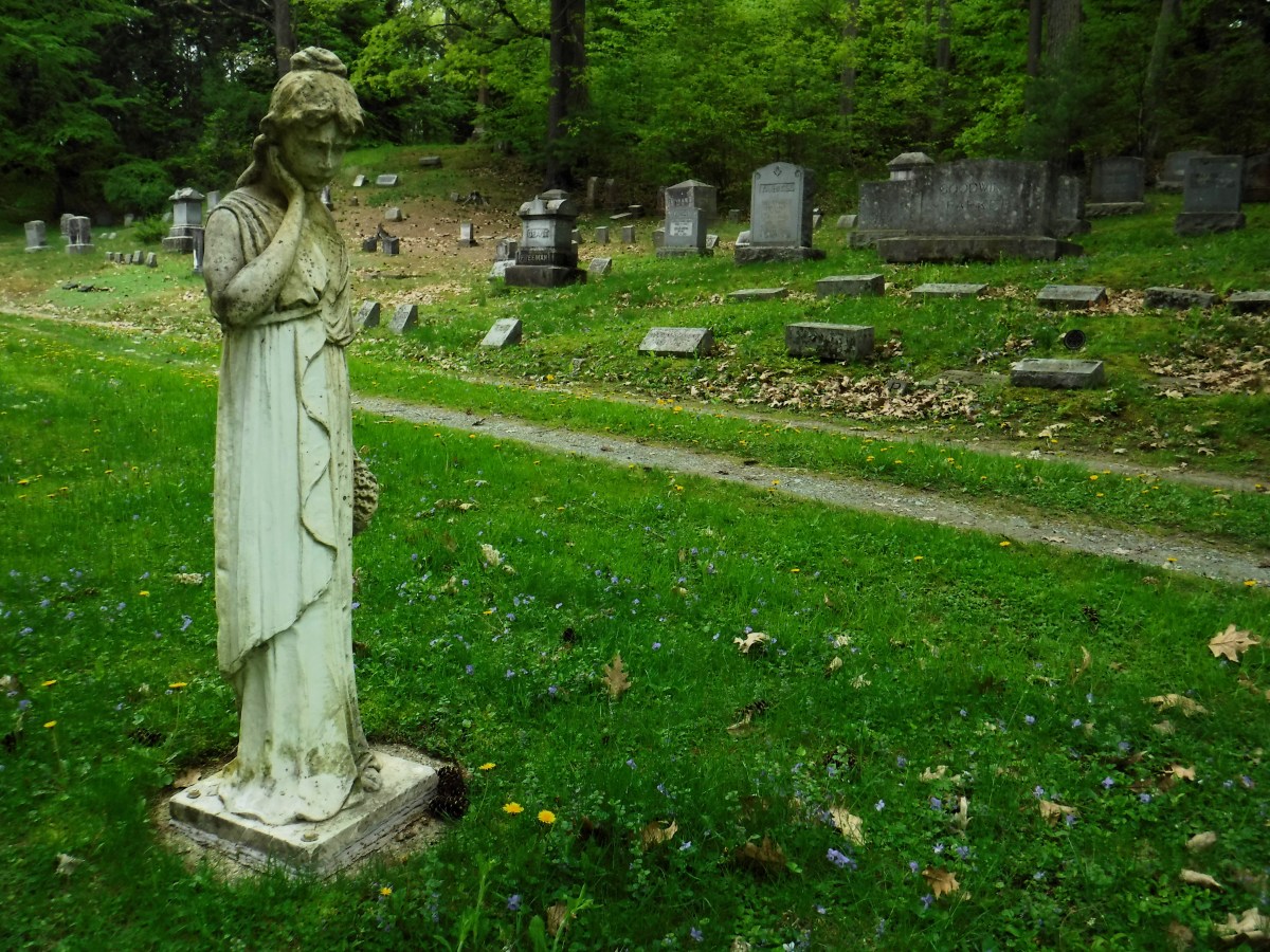 A weathered statue of a woman stands in a green cemetery with tombstones in the background on a cloudy day.