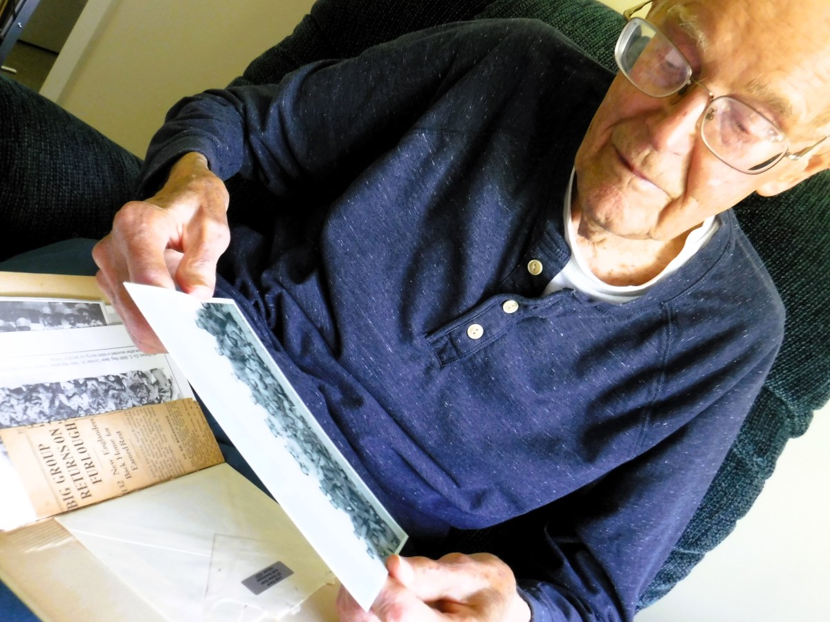 Elderly man wearing glasses and a navy blue shirt looks at an old photograph while reading a book on his lap, sitting in a dark green armchair.