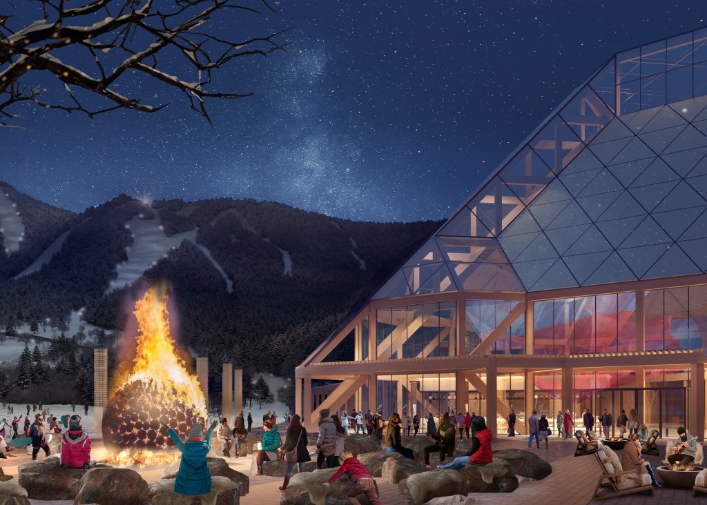 People gathered around a large bonfire outside a modern, glass-walled building at night, with snowy mountains and a starry sky in the background.