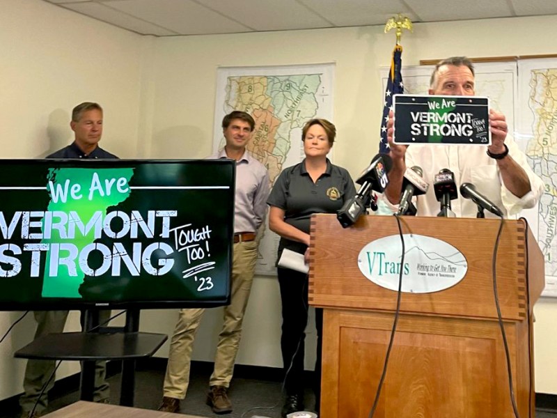 A man holds up a "We Are Vermont Strong" license plate at a podium in front of other people and a TV with the same message.