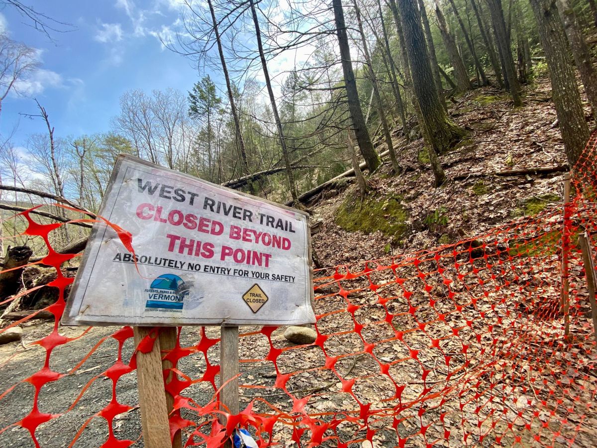 A weathered sign reads "west river trail closed beyond this point" beside a red safety net, in a wooded area.