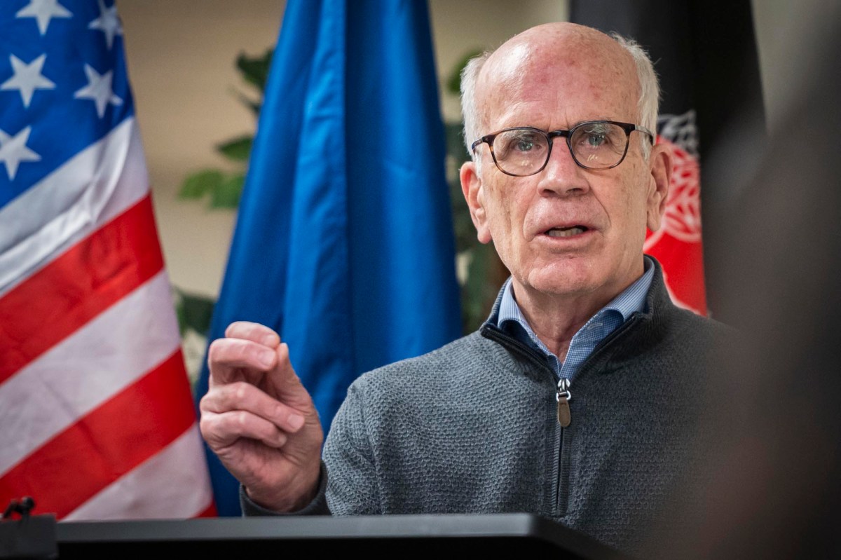 A man with glasses and wearing a gray sweater speaks at a podium. Behind him are flags.