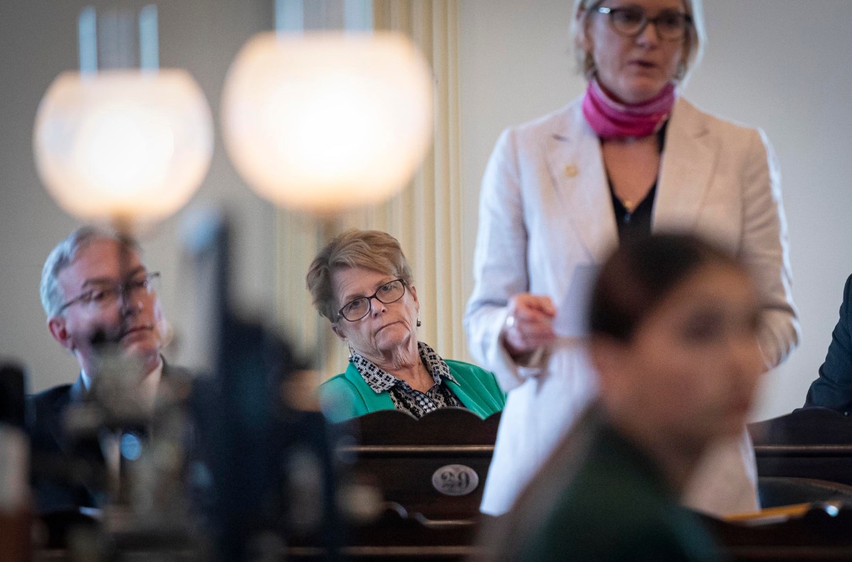 Focused woman with glasses listening intently in a meeting, background blurred with other attendees and foreground showing lamp silhouettes.