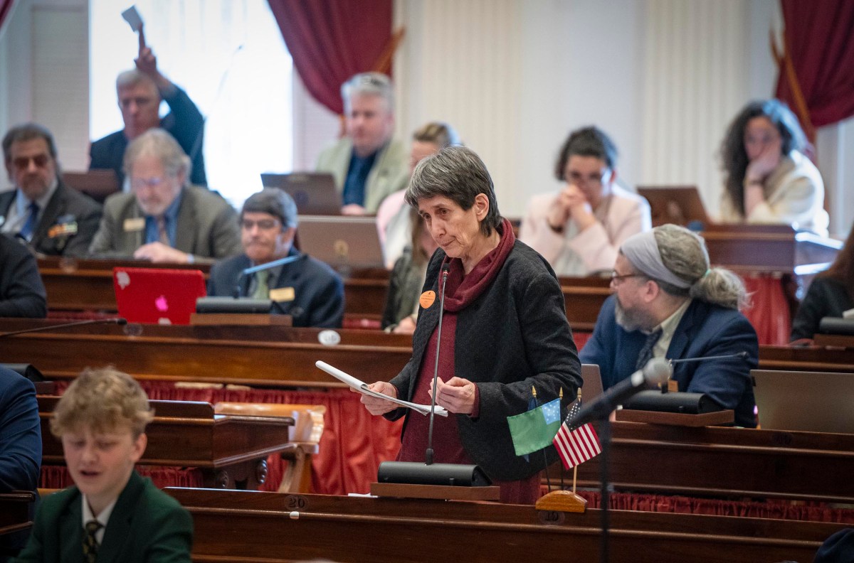 A legislator reads from papers in a busy assembly chamber, while other attendees are engaged in various activities.