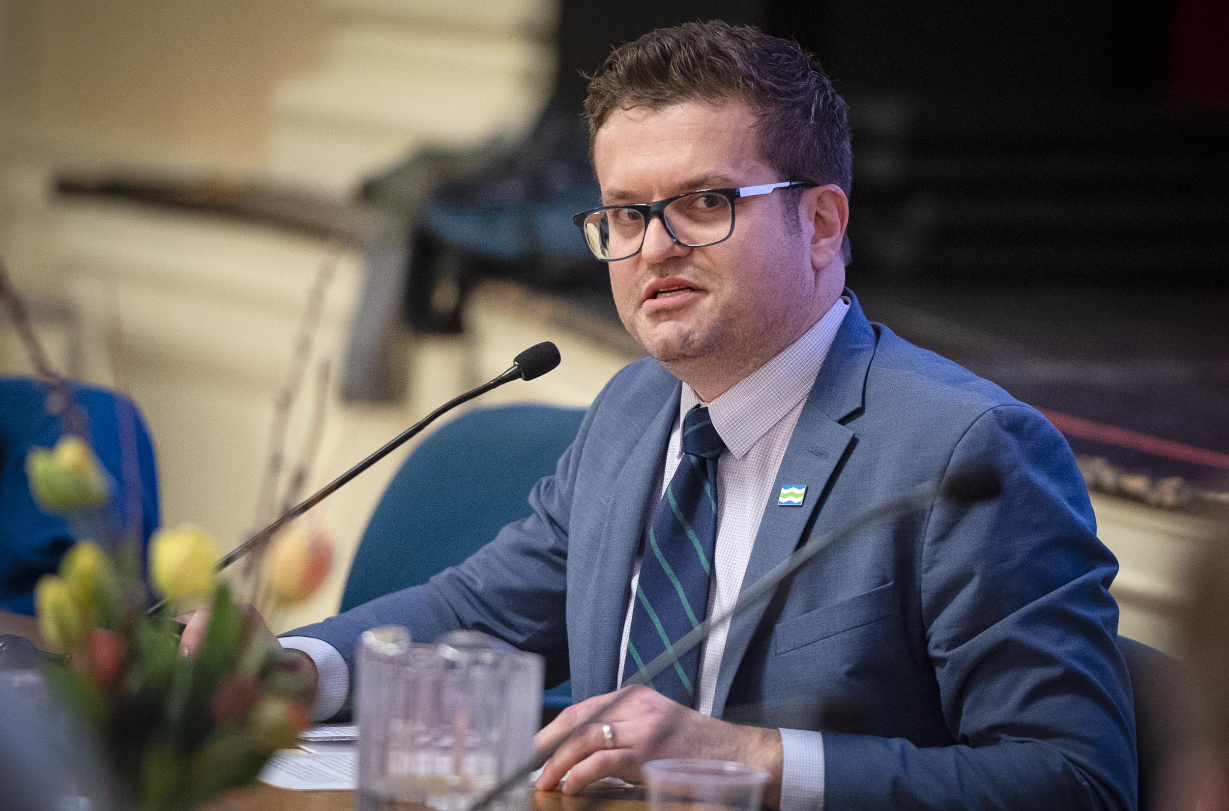 A man with glasses speaking at a conference, with a microphone in front of him and a blurred background.