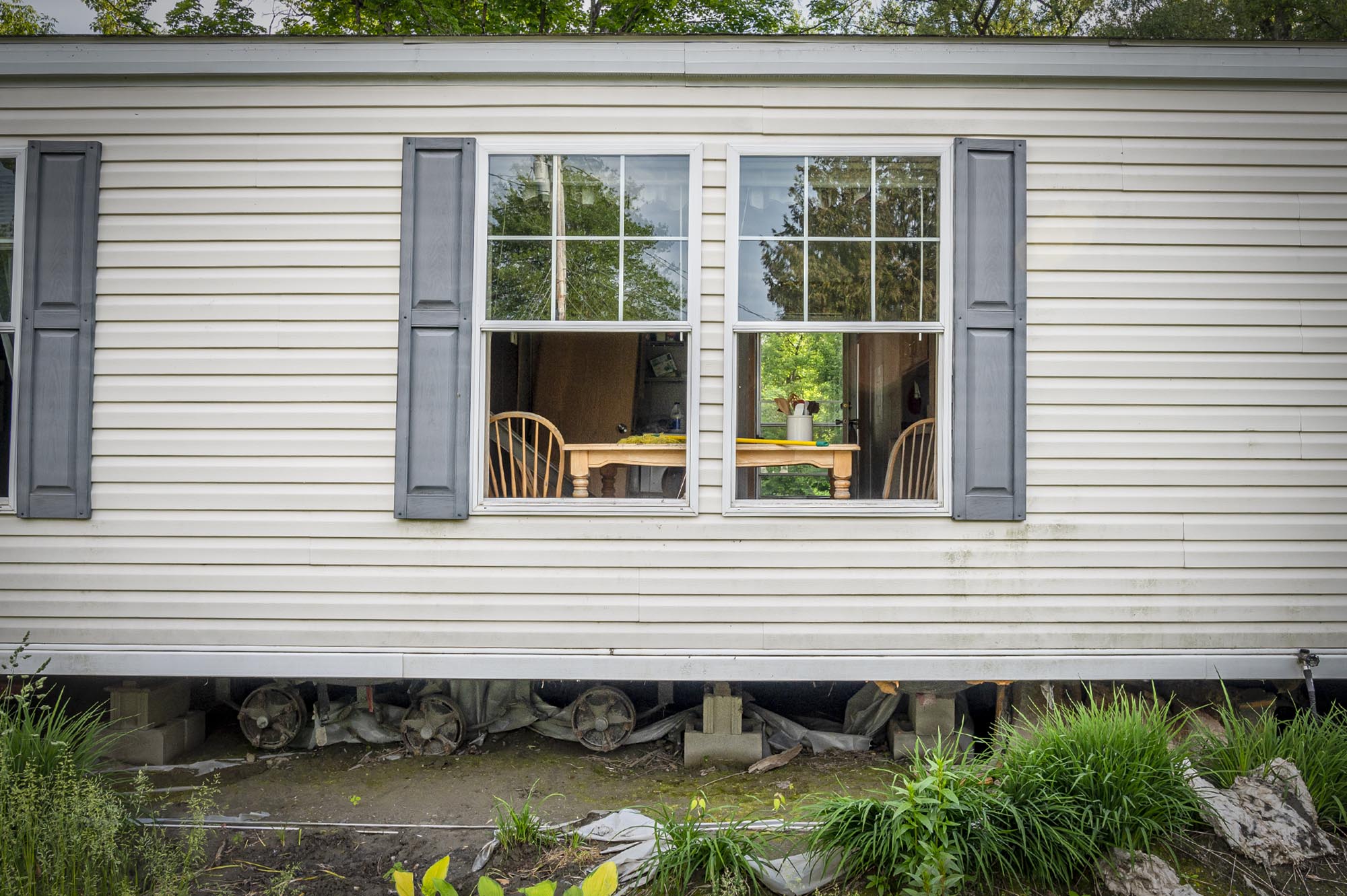 A white mobile home with two open windows, displaying a table and chairs inside. The home's exterior includes shutters and visible foundational supports beneath.