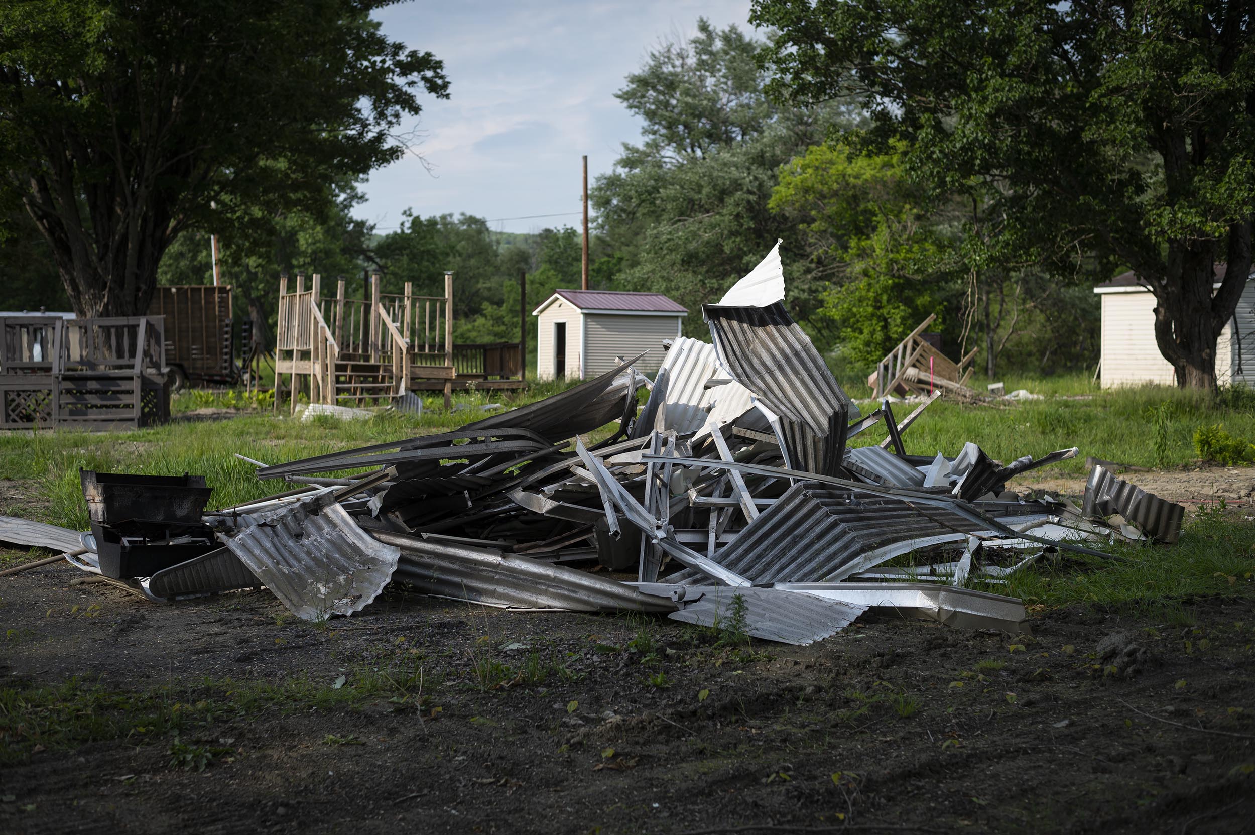 A pile of discarded corrugated metal sheets and debris is situated on the ground in front of trees and a small storage shed.