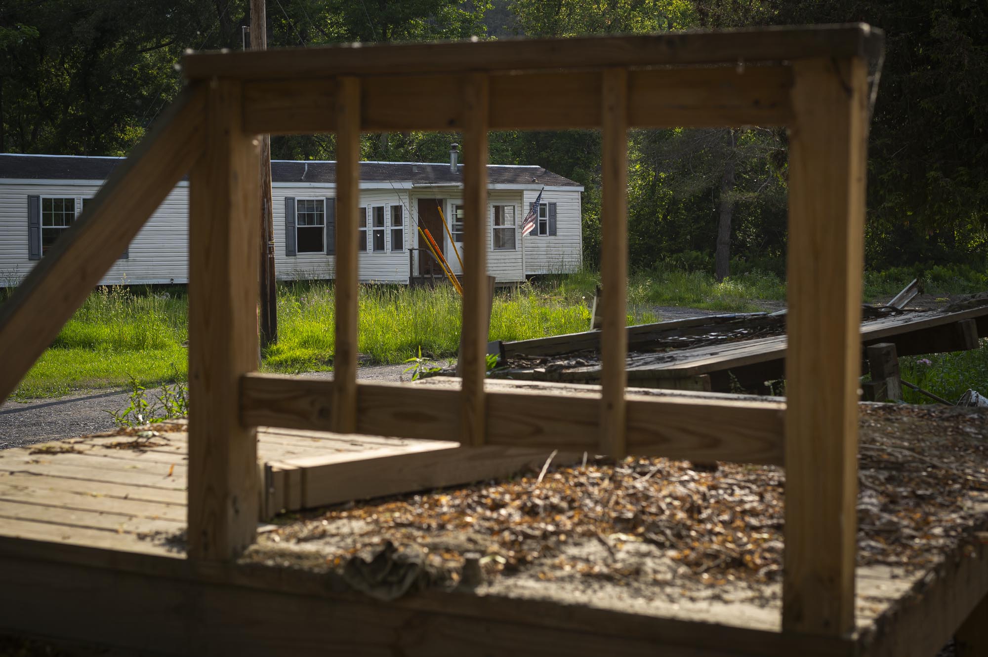 A mobile home is seen in the background through the wooden frame of a structure in the foreground, with overgrown grass and scattered debris around the area.